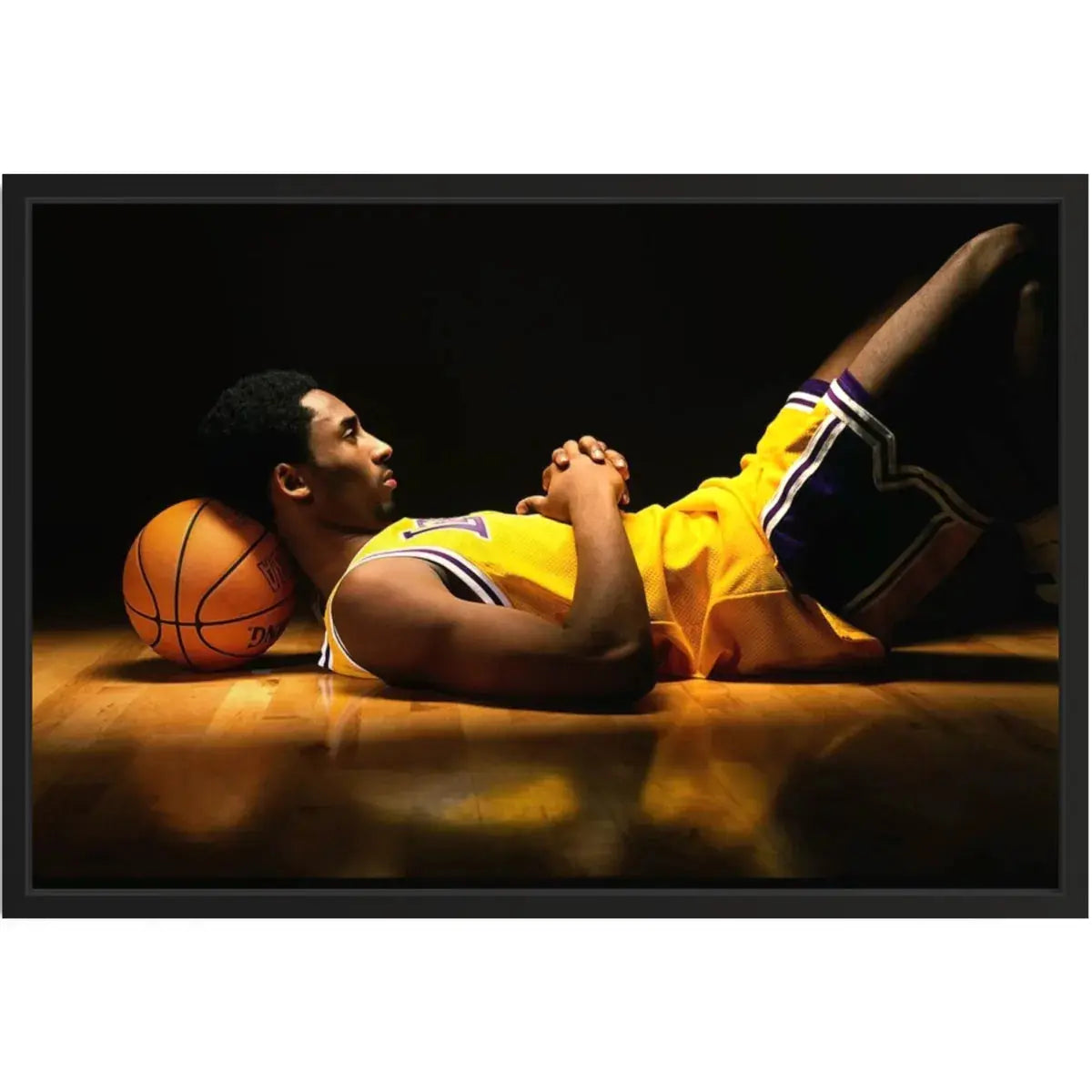 Close framed product shot of a basketball player in a yellow jersey resting on the court with a ball under his head on a black backdrop.