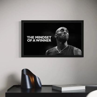 Framed poster with a black and white image of a person and text 'The Mindset of a Winner' on a wall above a desk.