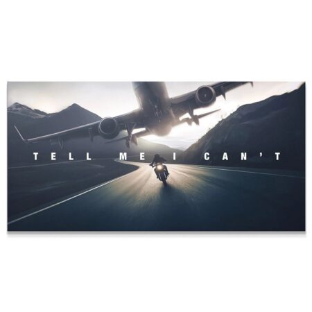 Wide shot of the motorcycle rider on the winding road below the airplane expressing "Tell Me I Can't" across the sky.