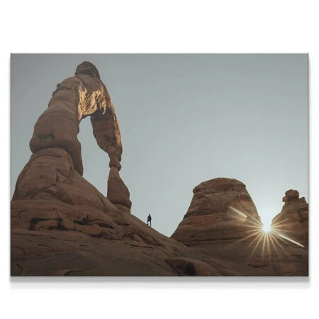 Low-angle view of 'The Arch Walker' highlighting the colossal scale of the sandstone arch against a pale sky.